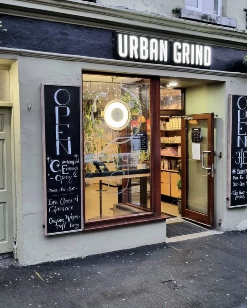The Urban Grind shopfront by day, with chalkboards either side of the open door and plants hanging above.
