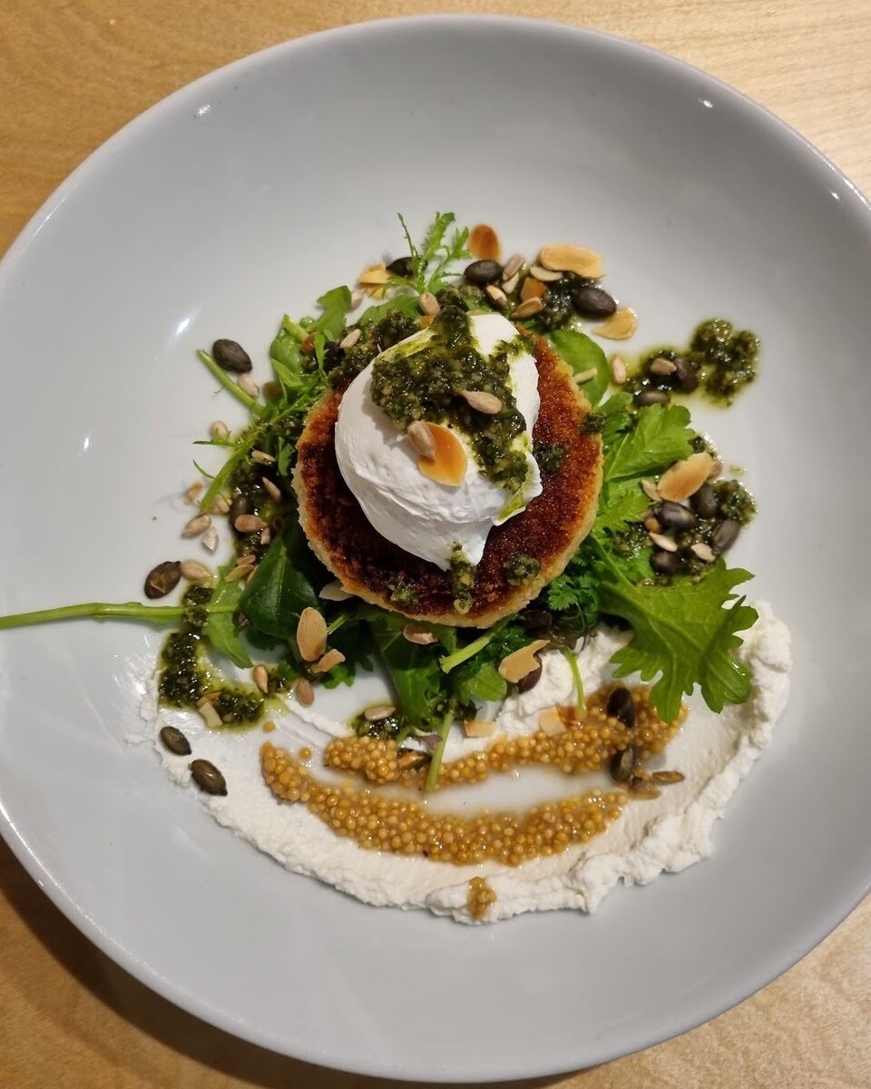 A poached egg on a crisp cake with greens, seeds, pesto and whipped cheese, in a white bowl on a wooden table.